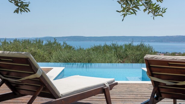 Eine Holzterrasse mit zwei Liegestühlen, die auf einen klaren, blauen Pool und Meerblick ausgerichtet ist.