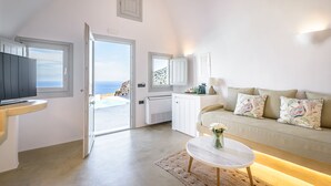 A modern living room with a sofa, a coffee table, and a TV. There is a door leading to a balcony with a view of the sea.