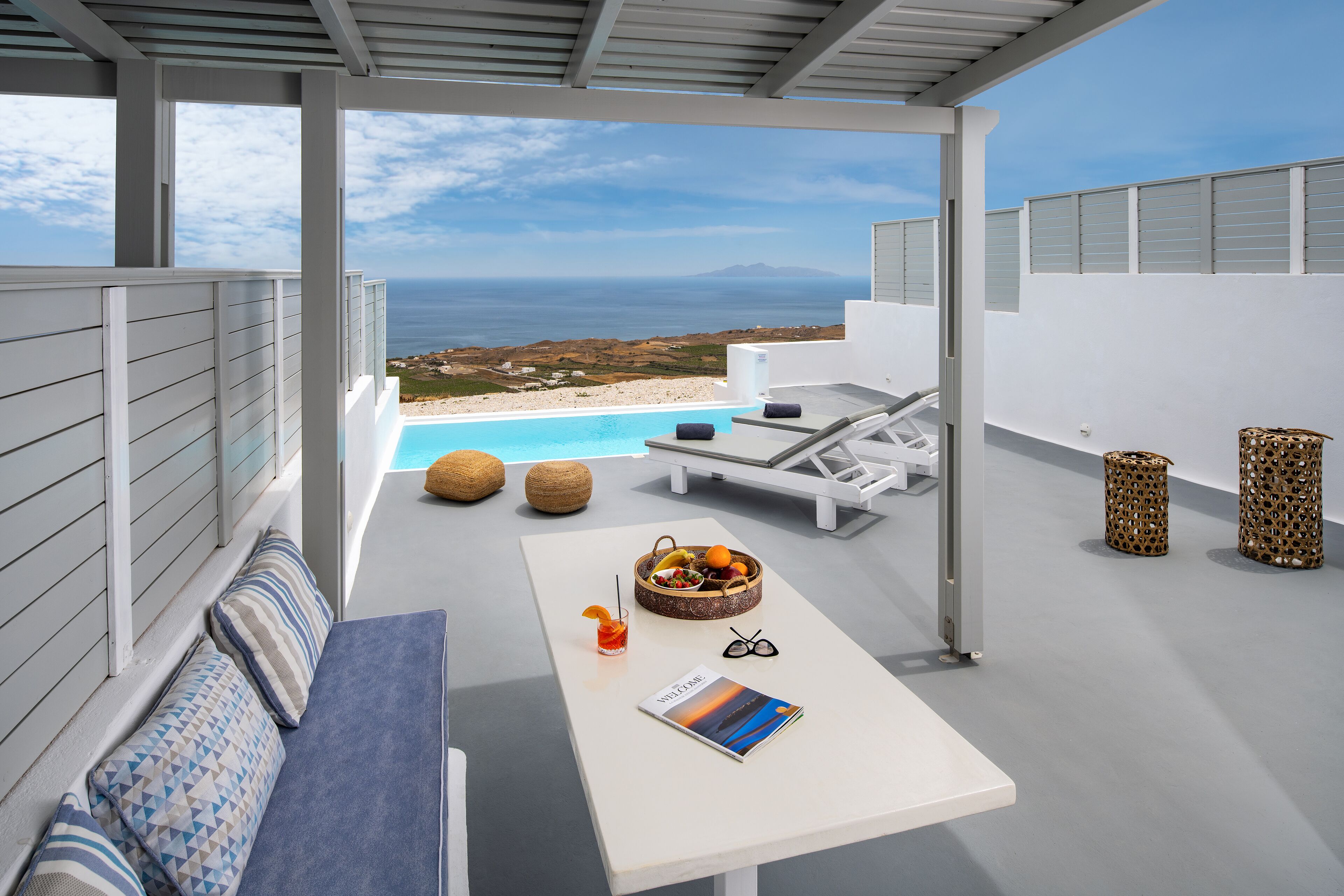 Uma área de estar moderna ao ar livre com piscina, uma mesa com bebida e revista, e vista para o mar.