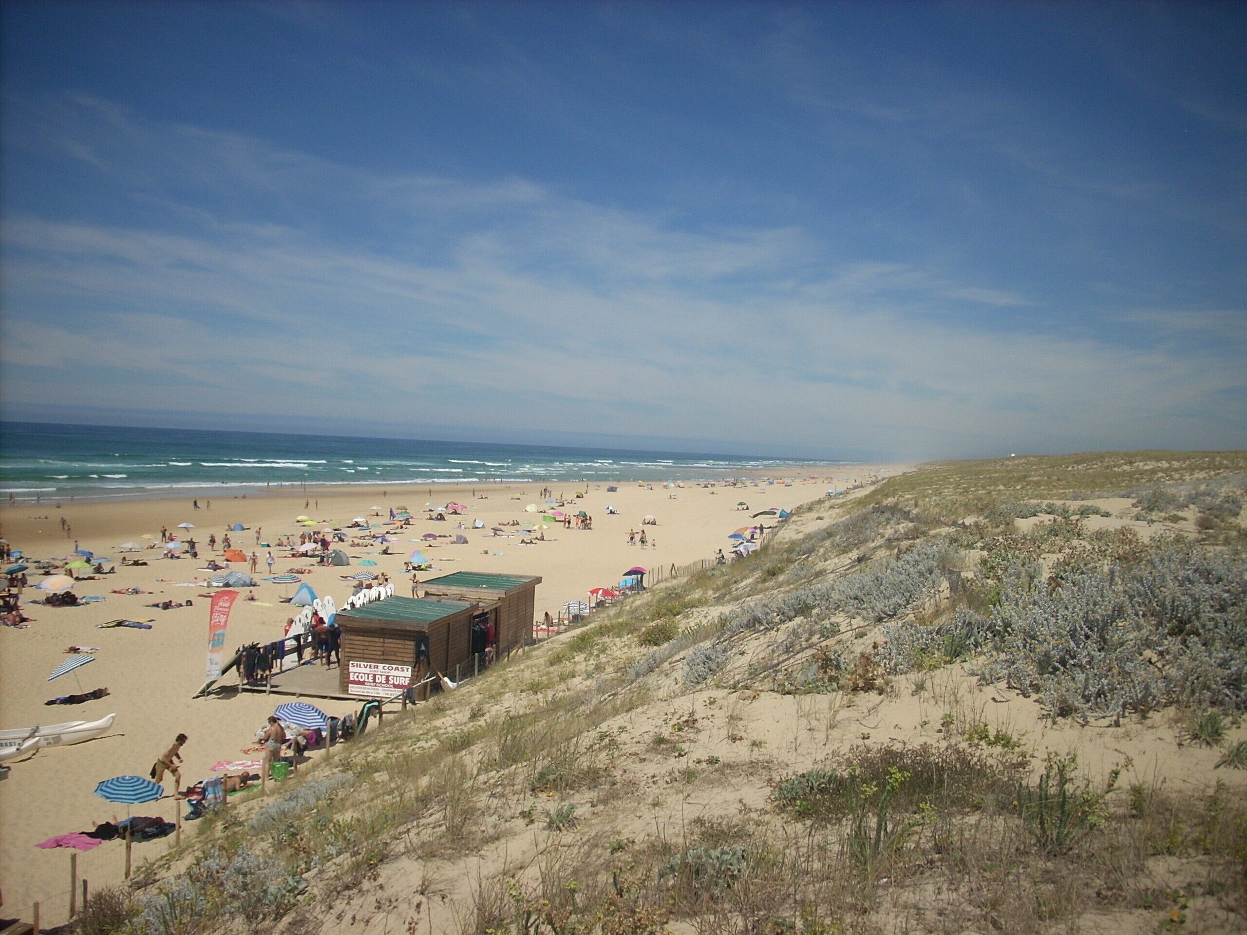 A beach with a sandy shore, a wooden structure labeled 'Ecole de Surf', and sparse vegetation on the dunes.
