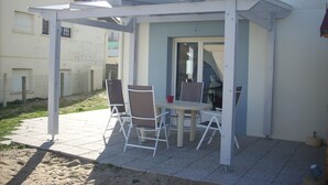An outdoor patio area with a table and chairs, a covered seating area, and a view of a residential building in the background. - Appart. 48m2 large garden terrace located 60m from the beach and 300m from the village (Mimizan Plage)