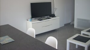 A modern, minimalistic room with a flat-screen TV on a white cabinet, a dining table with chairs, and a tiled floor. - Appart. 48m2 large garden terrace located 60m from the beach and 300m from the village (Mimizan Plage)