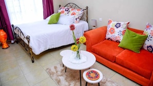 A bedroom with a bed, a sofa, a small table with a vase of flowers, and a colorful wall hanging. - Sherry Homes West suites (Nairobi)