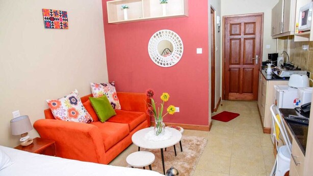 A compact living space with a red sofa, a small round table with flowers, a white round table, and a kitchenette area. - Sherry Homes West suites (Nairobi)