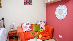 A cozy room with a red wall, a round mirror, a sofa with colorful pillows, a small table with a vase of flowers, and a nightstand with a lamp. - Sherry Homes West suites (Nairobi)