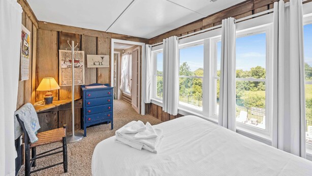 Een slaapkamer met een groot bed, een bureau met lamp, een stoel, een dressoir en uitzicht op groen door grote ramen.