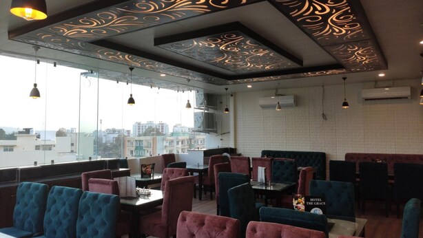 A restaurant with high tables and cushioned chairs, large windows with a city view, and decorative ceiling patterns. - Hotel The Grace (Rishikesh)