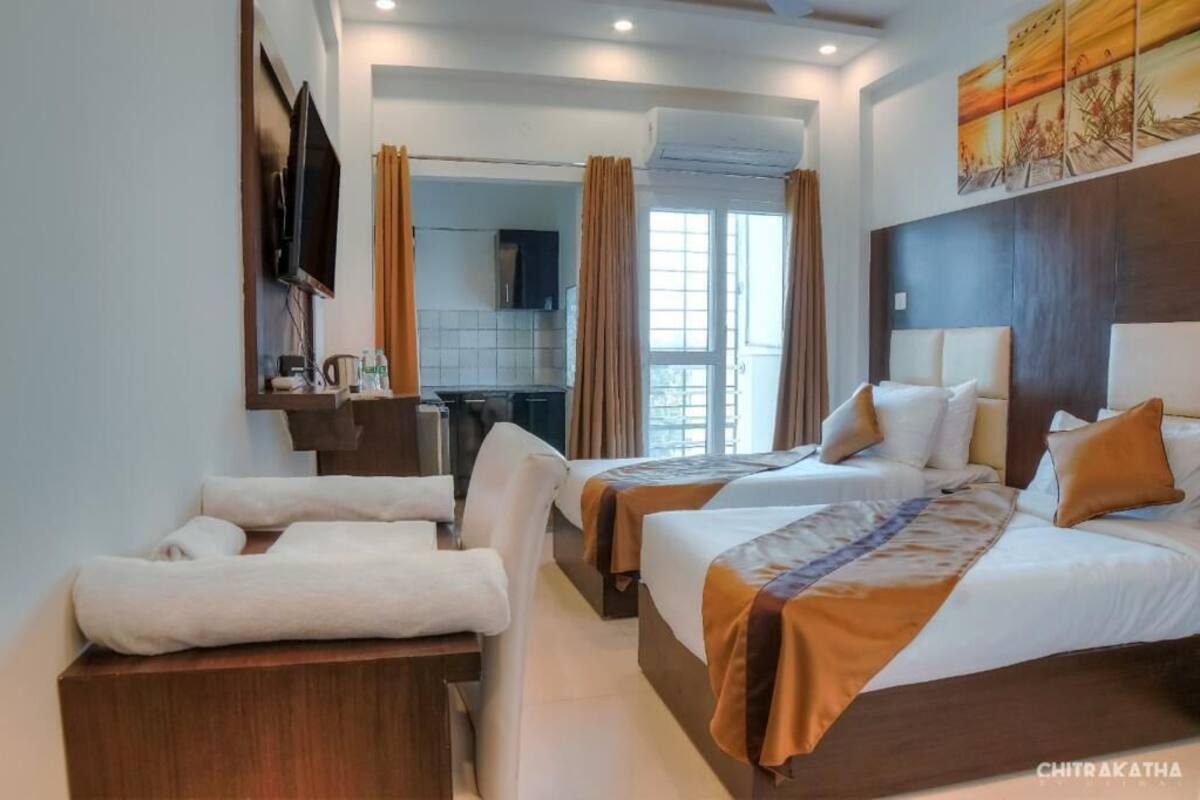 A hotel room with two beds, a desk, a chair, a TV, and a view of the kitchen and outside.