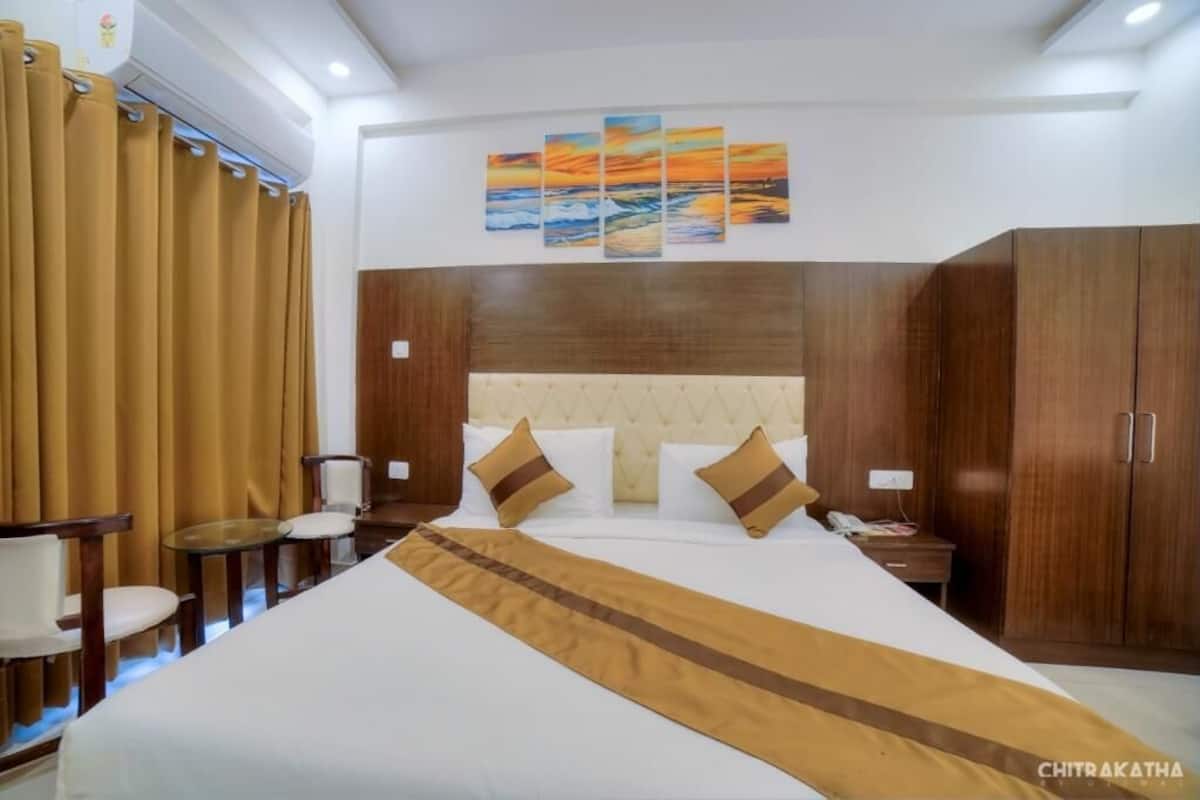 A hotel room with a large bed, a headboard with a wooden panel, a bedside table with a phone, and a wall with a sunset painting.