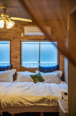 Ein Schlafzimmer mit Backsteinwand, zwei Fenstern, einem Deckenventilator und einem Bett mit weißer Bettwäsche und Kissen.