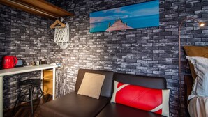 Ein modernes Zimmer mit Backsteinwand, einem Gemälde eines Piers, einer Couch mit Kissen, einem Schreibtisch mit rotem Wasserkocher und einer Stehlampe.