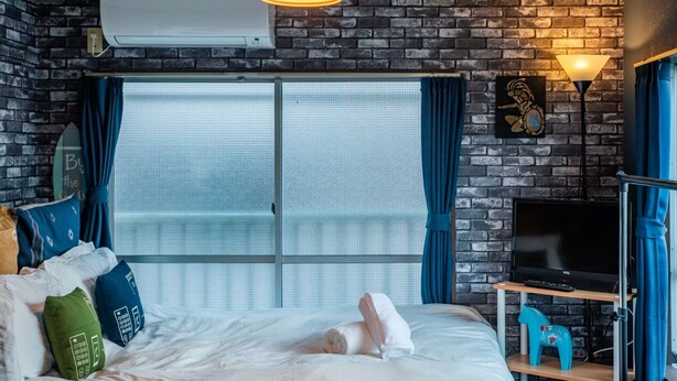 Ein Hotelzimmer mit Backsteinwand, großem Fenster, einem Bett mit Kissen, einem Fernseher auf einem Schrank und einem Deckenventilator.