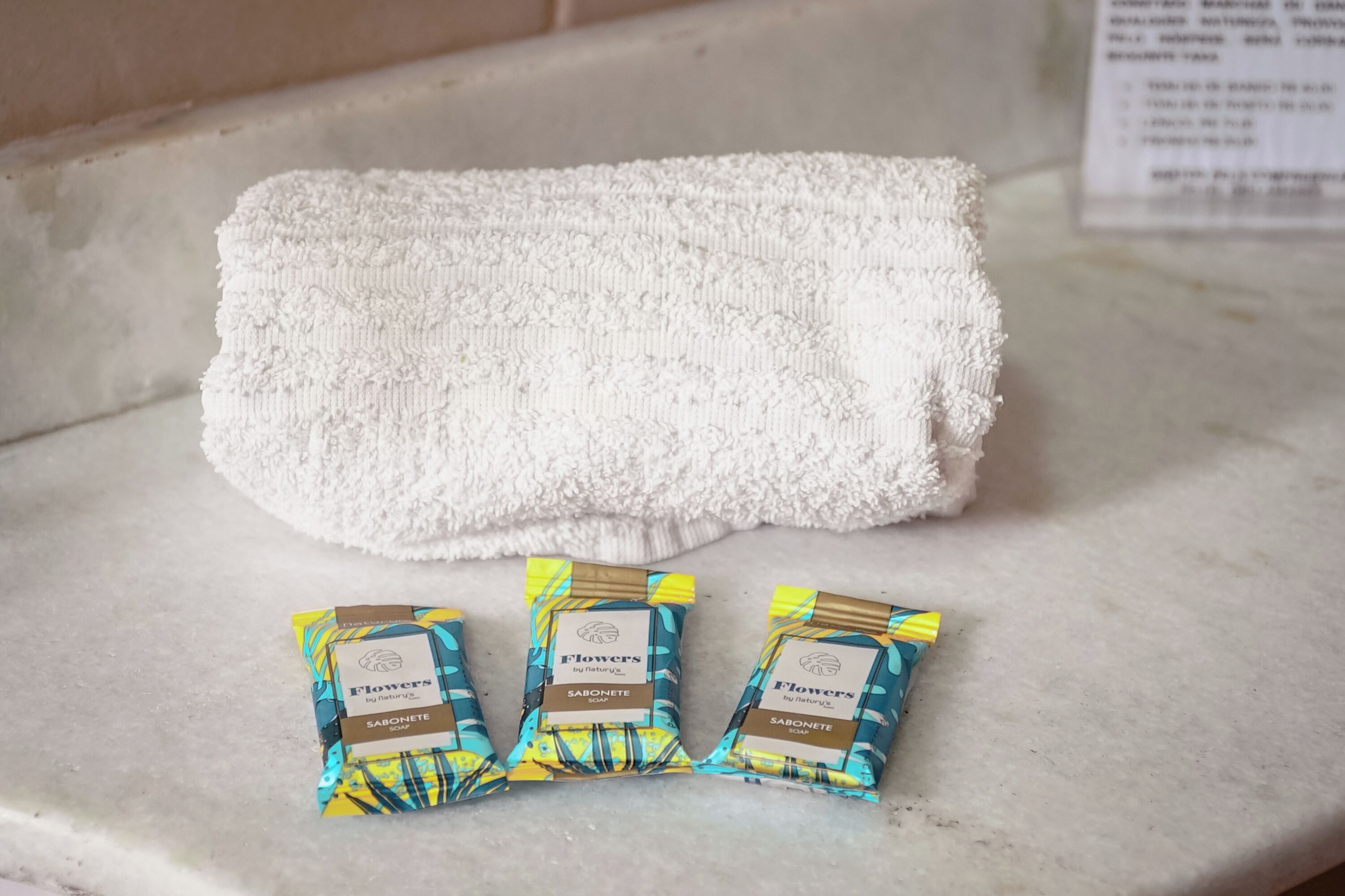 A bathroom with a white towel and three packets of Flowers by Nature soap.
