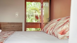 A bedroom with a wooden headboard, a bed with a floral-patterned pillow, and a view of palm trees through an open door. - Hotel Dellamares (Ubatuba)