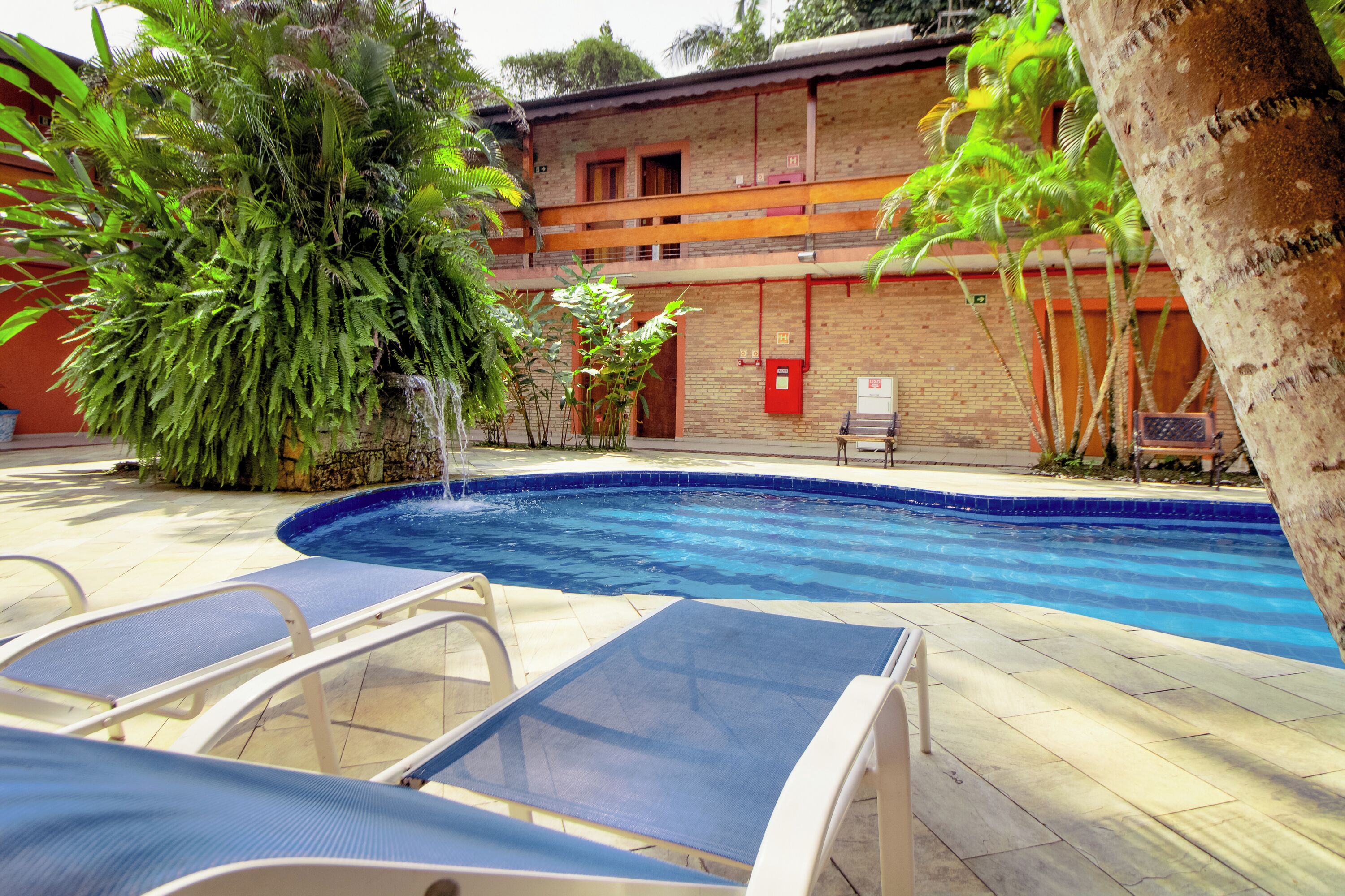 Una piscina con sillas de descanso, rodeada de abundante vegetación y un edificio de ladrillo con un buzón rojo.