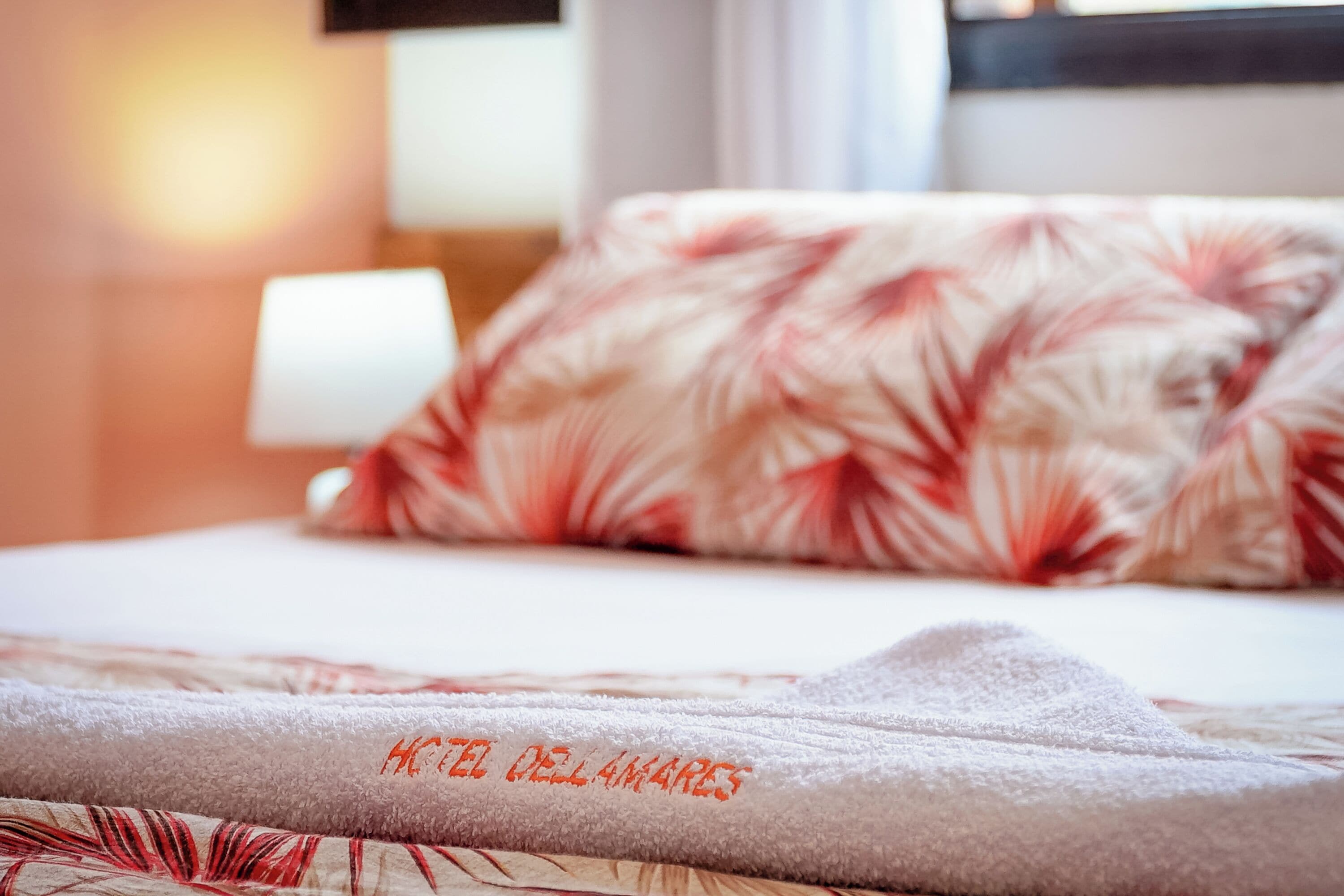 A hotel bed with a floral-patterned pillow and a towel with "HOTEL DELAMARES" embroidered on it.