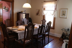 A dining room with a table set for six, surrounded by wooden chairs. A cabinet with decorative items is against the wall, and a window with curtains is visible.