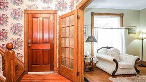 Un escalier en bois mène à une porte ornée d’un papier peint à motifs floraux, près duquel se trouvent un fauteuil blanc et une lampe.