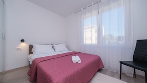 Un lit bien fait, recouvert d’une couverture rose, une pile de serviettes enroulées et une chaise noire dans une pièce lumineuse, dotée d’une fenêtre et d’une lampe fixée au mur.