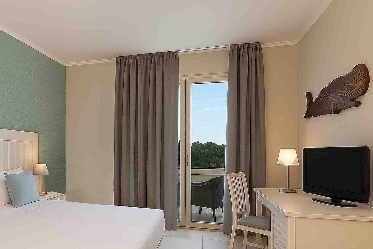 A hotel room with a bed, a desk with a TV, a chair, and a balcony with curtains.