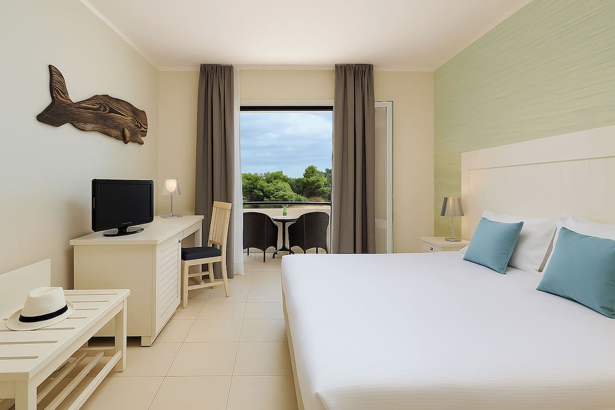A hotel room with a bed, a desk, a television, and a balcony with a view of trees.