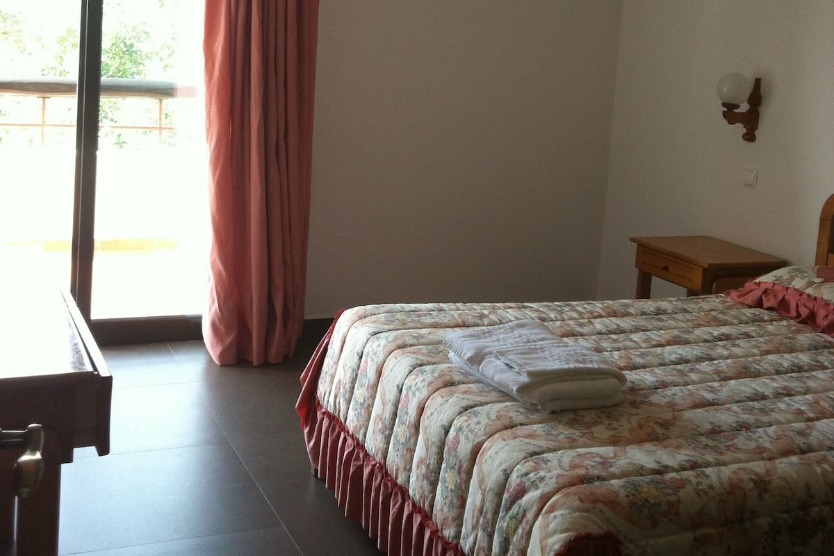 A bedroom with a bed, a desk, a chair, a window with curtains, and an air conditioning unit.