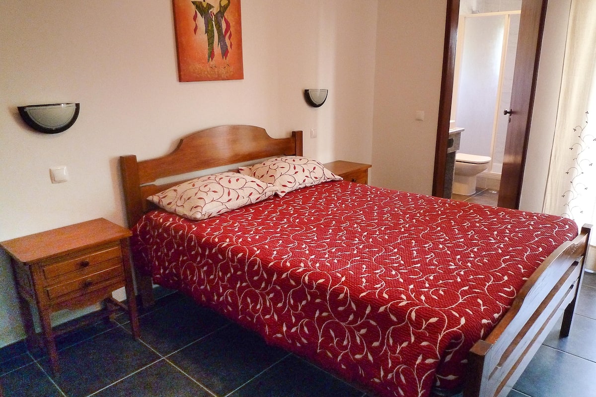 A bedroom with a bed, wooden nightstands, a red patterned bedspread, a wall-mounted artwork, and a bathroom visible through an open door.