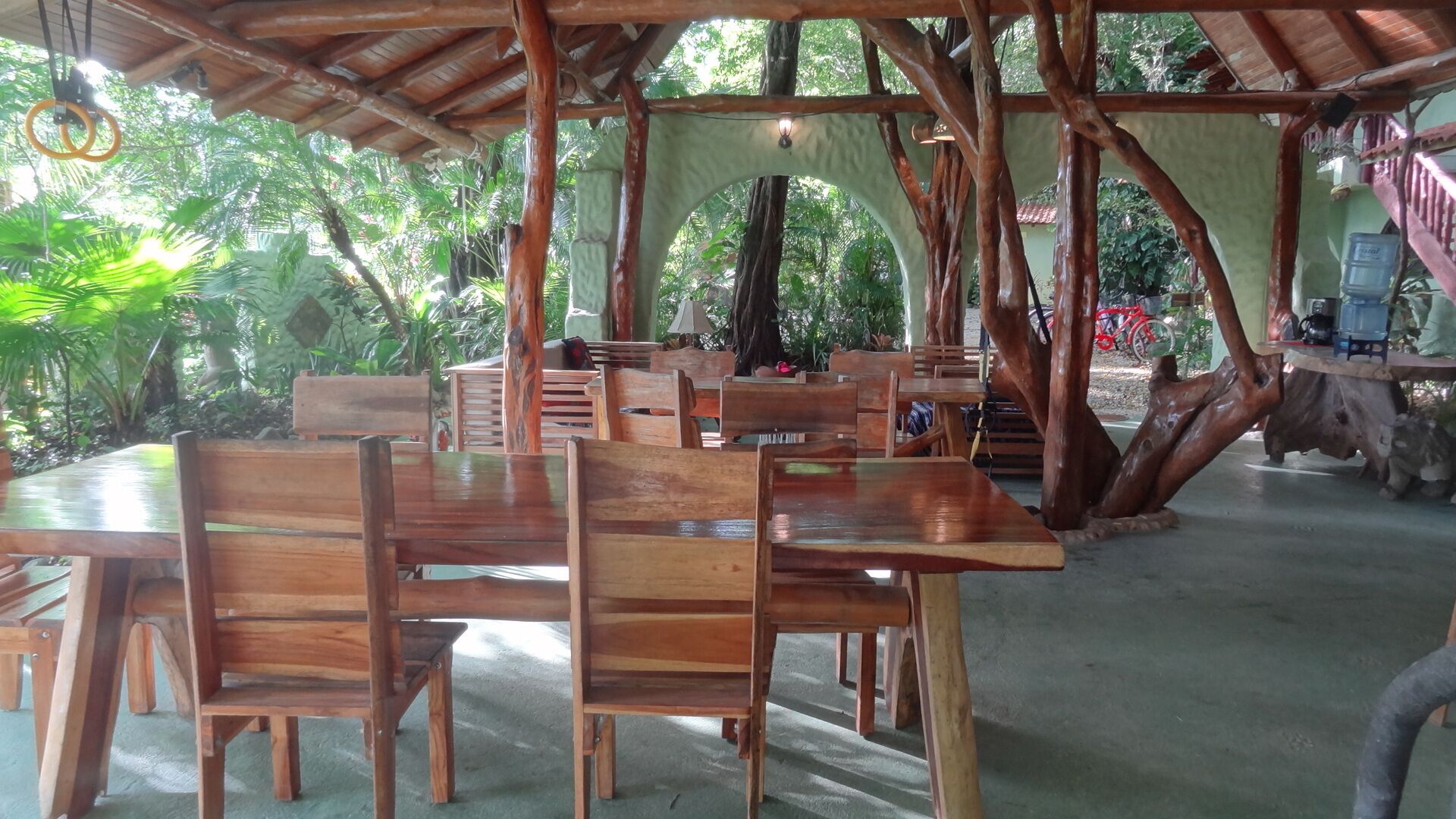 Een terras buiten met houten eettafels en stoelen, omringd door bomen en een overdekte constructie.