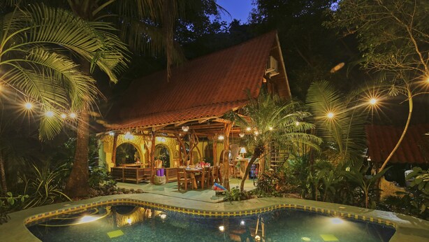 Un área junto a la piscina con una estructura de madera, una piscina y vegetación tropical circundante.