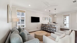 Una sala de estar moderna con un sofá gris, un sofá blanco, una zona de cocina con barra y un ventanal grande.