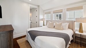 Un dormitorio con una cama grande, una mesita de noche de madera, una lámpara y vistas al exterior a través de las ventanas.
