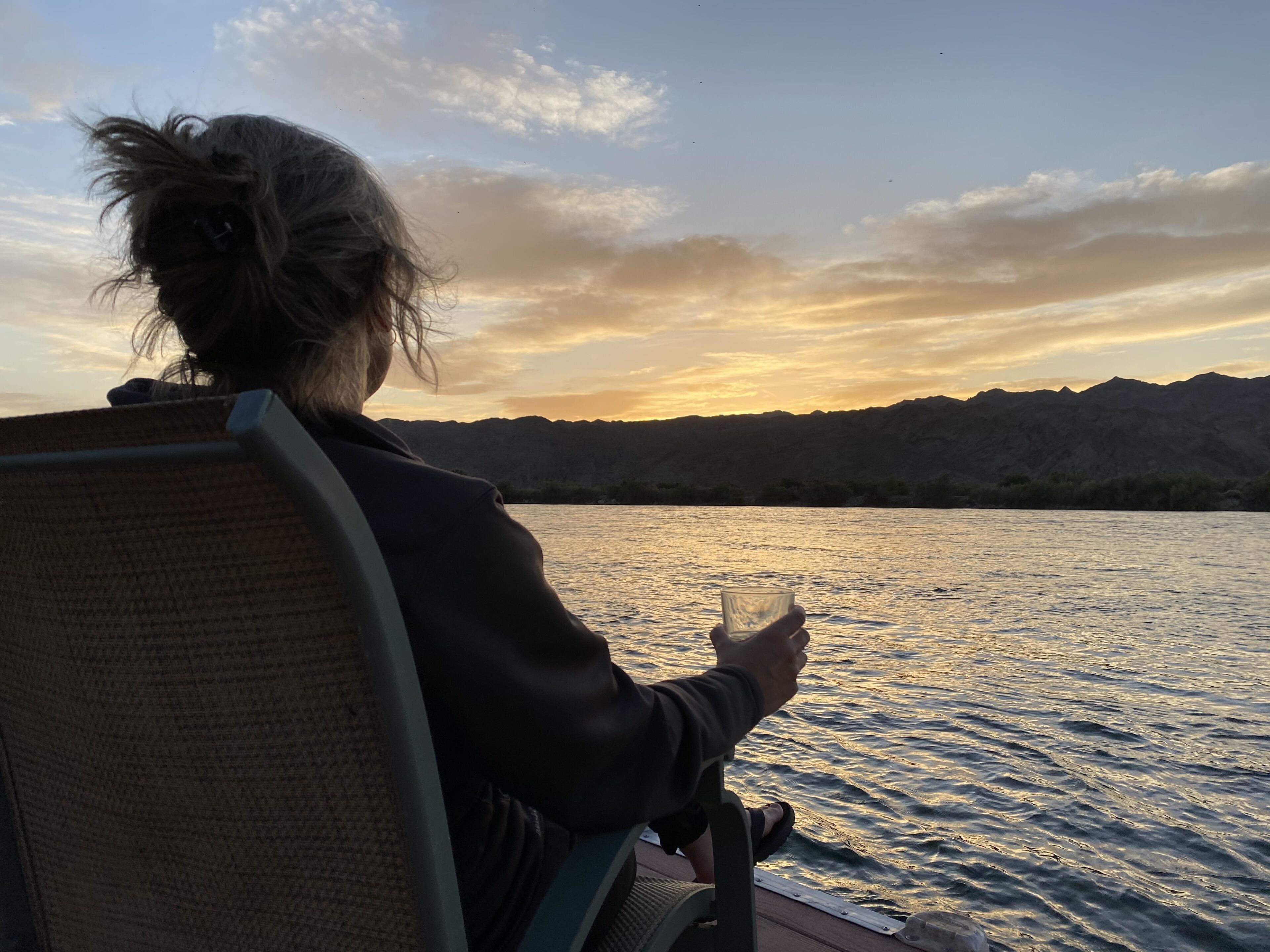 A person seated by a body of water during sunset, with mountains in the background.