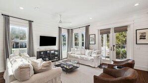 Amplia sala de estar con un sofá grande, una mesa de centro y un sillón de cuero. Hay un televisor de pantalla plana montado en la pared, y la habitación tiene ventanas grandes con cortinas.