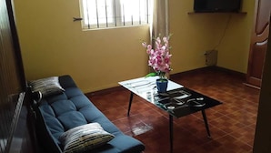 Ein Wohnzimmer mit einem dunkelblauen Sofa, einem kleinen schwarzen Tisch mit einer Vase voller rosa Blumen und einem an der Wand montierten Fernseher.