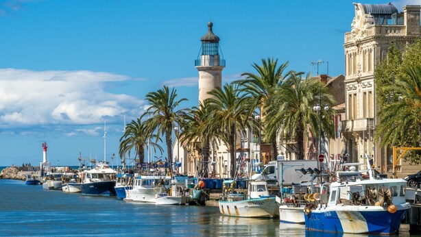 Un port où sont amarrés des bateaux, un phare et des palmiers qui bordent la promenade.