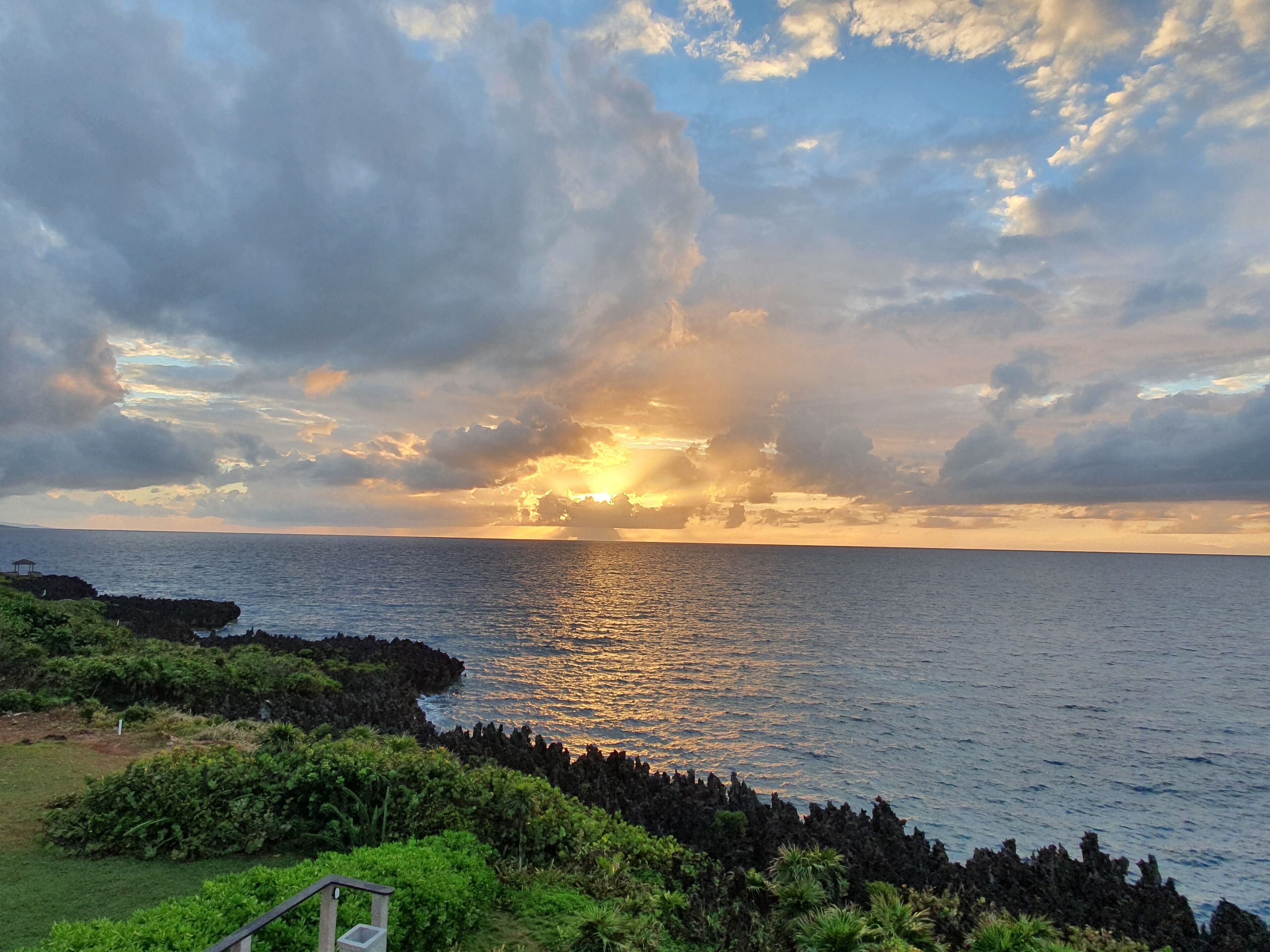 Paisagem costeira com um pôr do sol sobre o oceano, costa rochosa e vegetação exuberante.