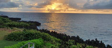 Paisagem costeira com um pôr do sol sobre o oceano, costa rochosa e vegetação exuberante.