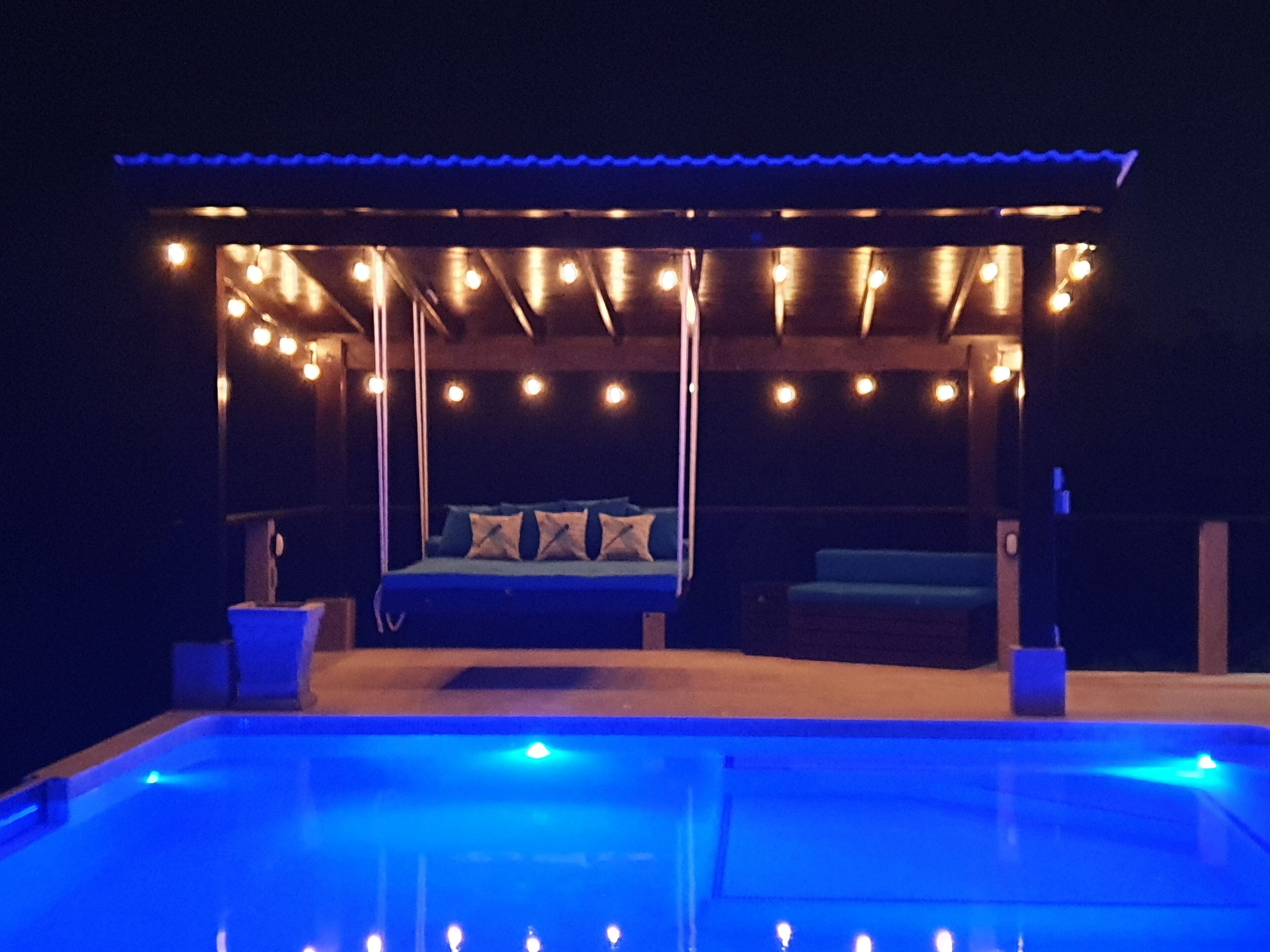 A pool area with a bed under a covered section, surrounded by string lights.
