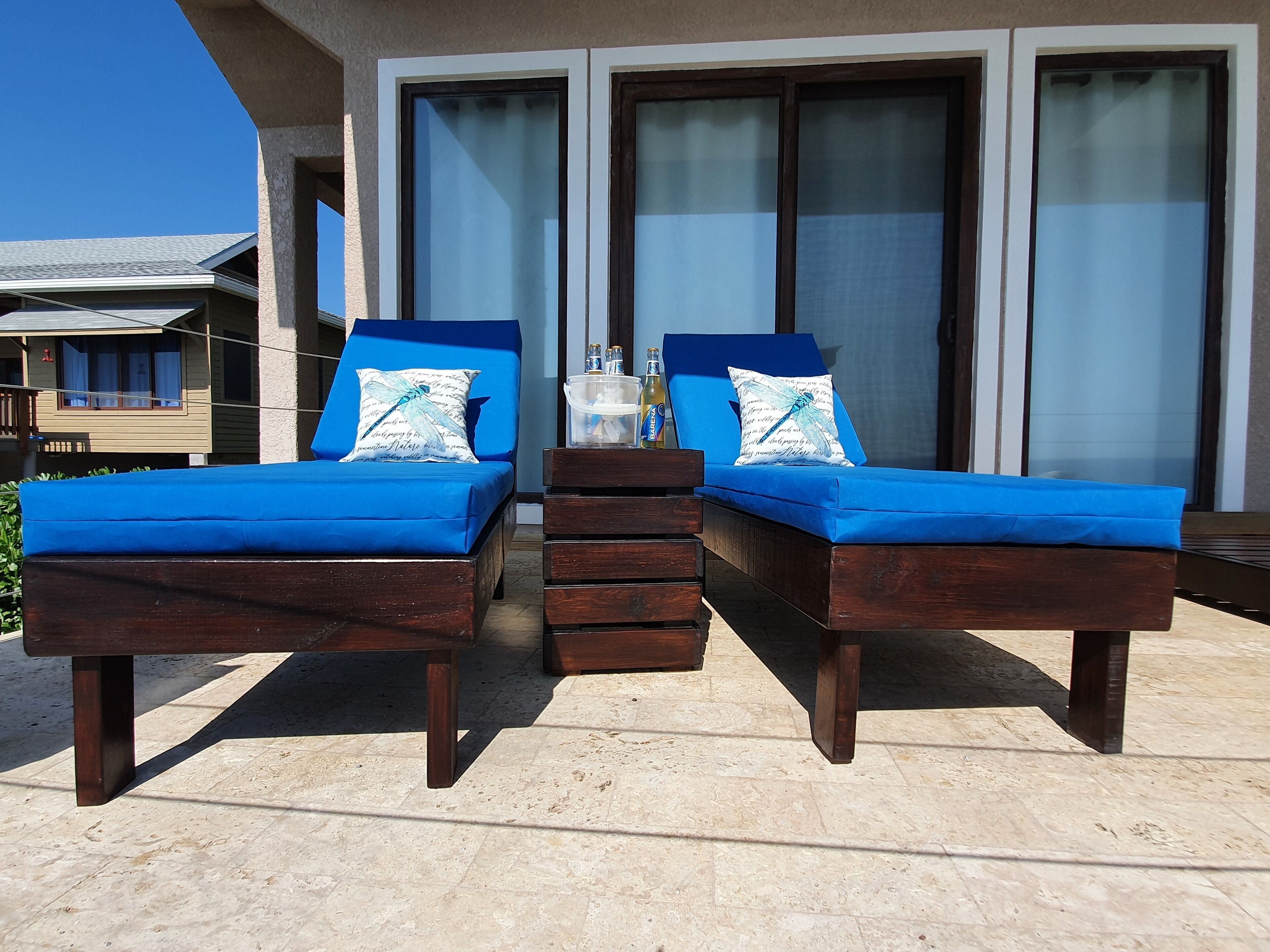 Two wooden lounge chairs with blue cushions and dragonfly-themed pillows, positioned on a tiled patio with a view of a building with large glass doors.