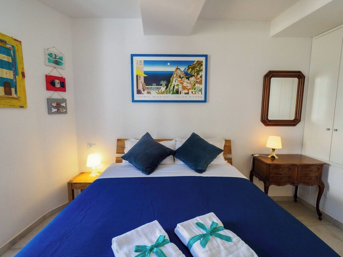A hotel room with a bed, pillows, a nightstand with a lamp, a dresser, a mirror, and a framed picture on the wall.