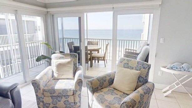 A balcony with two patterned armchairs, a dining table with chairs, and a small table with folded clothes. - Siesta Key Beach Front Condo! Breathtaking view! (Siesta Key)