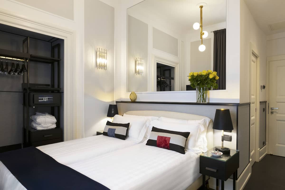 Ein Hotelzimmer mit einem ordentlich bezogenem Bett, einem Nachttisch mit Lampe, einem Spiegel über dem Bett und einem Regal mit Handtüchern und einer Blumenvase.