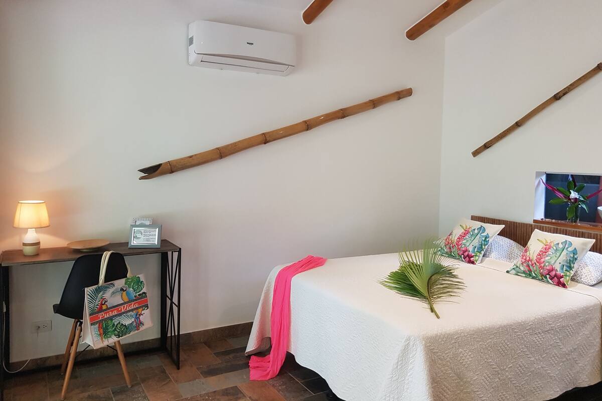 A bedroom with a bed, bedside table, lamp, air conditioner, and wooden beams on the ceiling.