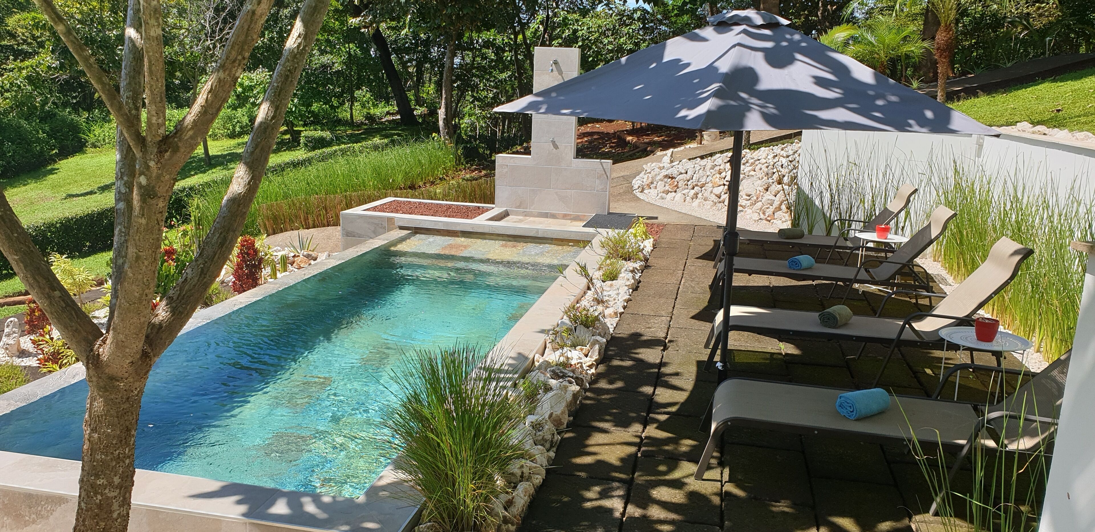 A rectangular swimming pool with a stone border, surrounded by a landscaped garden with trees and a patio area with outdoor furniture and a large umbrella.