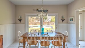 Une salle à manger avec une table en bois et des chaises blanches, un lustre, et une vue sur un jardin avec une piscine.