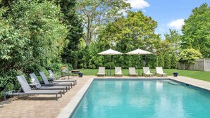 Une piscine rectangulaire dont le bord est recouvert de carreaux, entourée d’une terrasse en briques. Six chaises longues sont alignées le long du bord de la piscine, chacune étant accompagnée d’un parasol assorti. L’ensemble est bordé d’un feuillage verdoyant et dense ainsi que d’arbres.