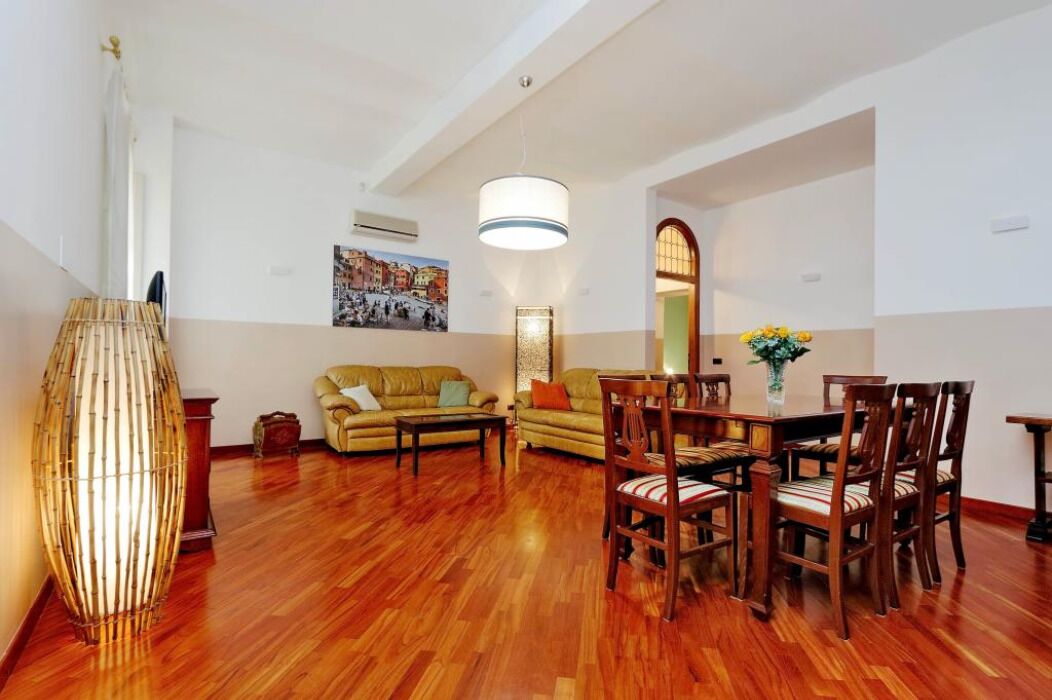 A spacious living room with a wooden floor, a sofa set, a dining table with chairs, a large decorative lamp, and a wall-mounted mirror.