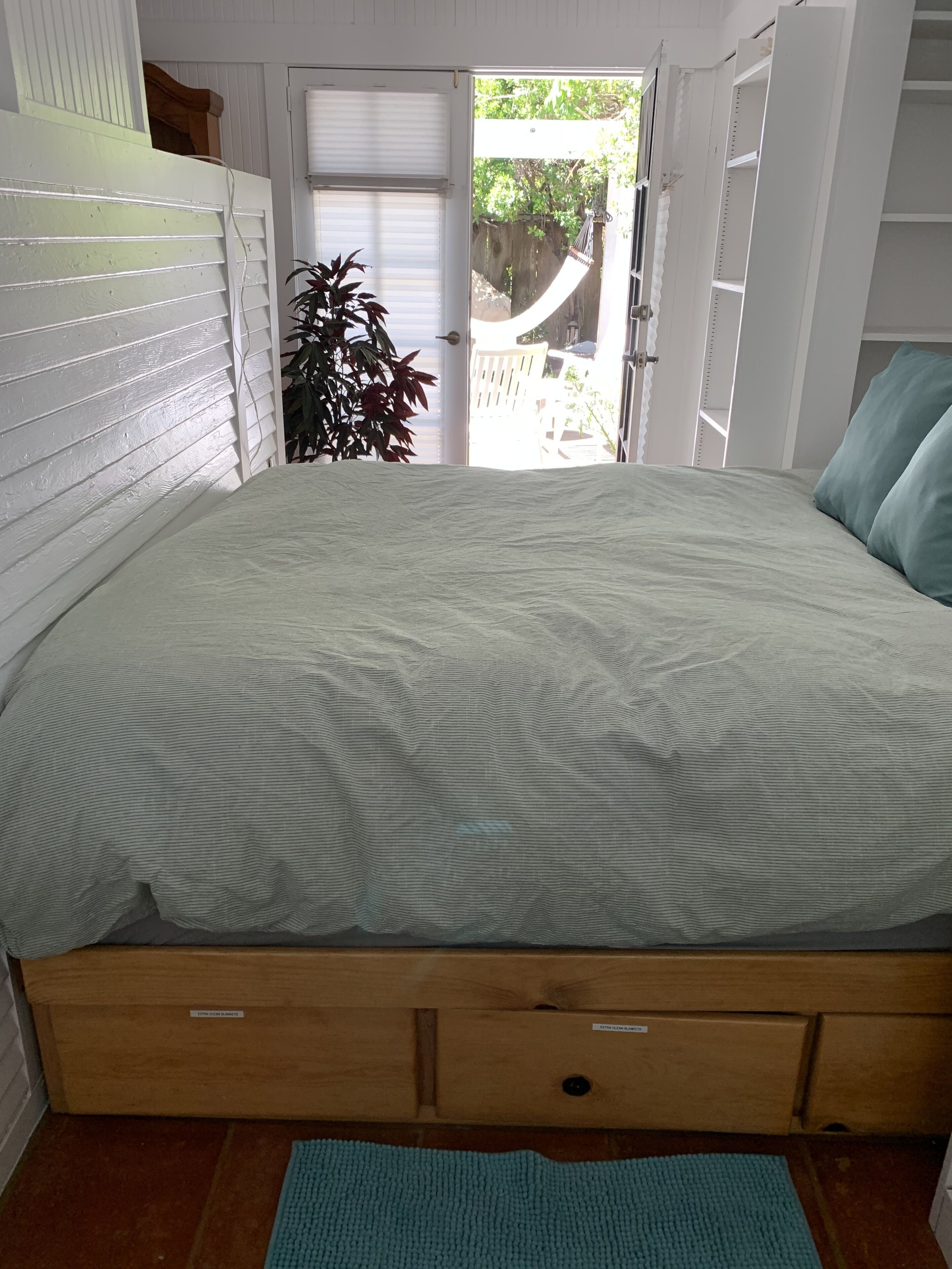 A bedroom with a bed, wooden drawers, a plant, and a view through an open door.