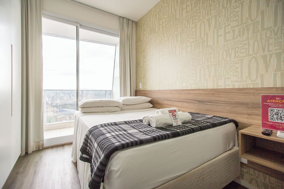 A hotel room with a large window, a bed with white linens, a check-in desk, and a QR code on the wall.