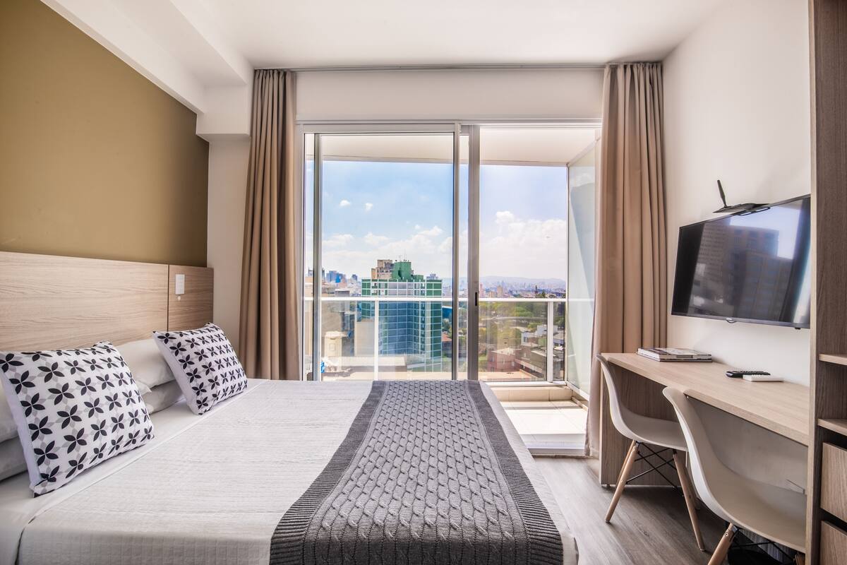 A hotel room with a large bed, a desk, a TV, and a balcony with a city view.
